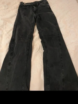 Svarta bootcut jeans med sned knäppning - Säljer ett par svarta bootcut jeans med cool sned knäppning i midjan. Jeansen har klassisk femficksdesign och är tillverkade i denim. Perfekta för dig som vill ha en unik twist på dina svarta jeans.