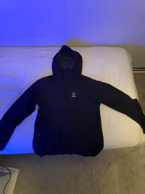 Svart regnjacka från Haglöfs med huva - Snygg svart regnjacka från Haglöfs med justerbar huva och dragkedja framtill. Jackan har meshfoder på insidan för extra ventilation och komfort. Perfekt för regniga dagar och outdoor-äventyr. Klassisk passform och minimalistisk design med diskret logga på bröstet. Kan gå ner i pris vid snabb affär