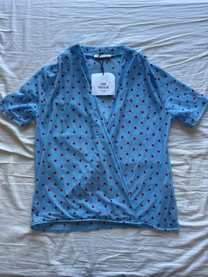 Blå omlottblus med röda prickar Zara - Snygg blå omlottblus från Zara Trafaluc med korta ärmar och röda prickar. Blusen har en djup omlottringning och är gjord i ett lätt, tunt material 