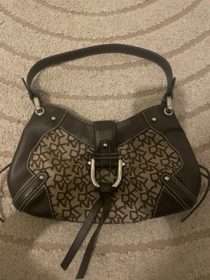 Brun DKNY handväska med monogram - Snygg handväska från DKNY i brunt läder och canvas med klassiskt DKNY-monogram. Väskan har silverfärgade metalldetaljer, dekorativt spänne framtill och kort band. Perfekt axelrem och cool form som ger en trendig look. Såååå snygg❤️❤️❤️ Använd gärna köp nu