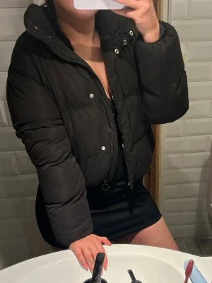 Svart kort pufferjacka - Säljer en svart, kort pufferjacka med hög krage och dragkedja framtill. Jackan har en boxig passform och är perfekt för kalla vinterdagar. Stängs med både dragkedja och tryckknappar. Enkel och stilren design som passar till det mesta.