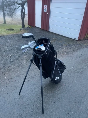 Golfbag med klubbor från Open - Säljer en svart och grå golfbag från Open med flera fack och bärremmar. I bagen ingår flera golfklubbor med silverfärgade huvuden. Perfekt för dig som vill komma igång med golf eller behöver extra utrustning.