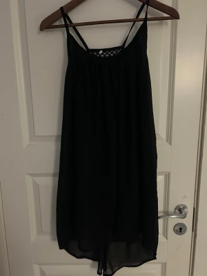 Svart ärmlös klänning med nät - Svart klänning med tunna axelband och ärmlös design. Framsidan har en snygg nätlik detalj upptill som ger en cool touch. Klänningen är luftig och har en avslappnad passform, perfekt för varma dagar. Materialet känns lätt och skönt mot huden.