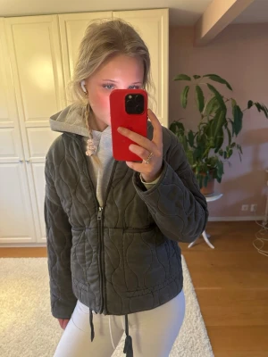 Vår jacka - Säljer en mörkgrå quiltad jacka med dragkedja framtill och huva. Jackan har ett snyggt mönster i quiltad stil och är långärmad. Perfekt för dig som vill ha en cool och bekväm look.