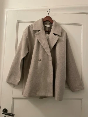 Beige ullkappa från ZARA - Stilren beige kappa från ZARA i ullblandning med klassisk krage och knäppning framtill. Kappan har en rak, loose passform och långa ärmar. Perfekt för dig som gillar minimalistisk och tidlös stil.