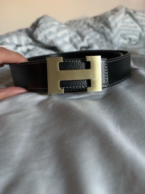 Svart skärp med guldspänne - Snyggt svart skärp med klassiskt H-format guldspänne. Skärpet är gjort i präglat skinn och har vita sömmar längs kanterna. Perfekt accessoar för att lyfta din outfit.