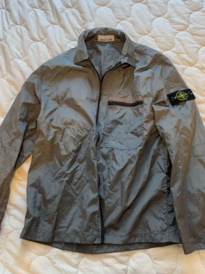 Grå overshirt från Stone Island - Snygg grå overshirt från Stone Island med dragkedja framtill och bröstficka med zip. Klassisk krage och ikonisk logotyp-patch på vänster ärm. Tillverkad i lätt, glansig nylon som ger en modern look.
