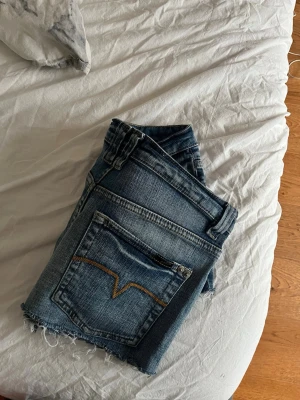 Blå jeansshorts med rå kant - Snygga blå jeansshorts med rå, fransig kant nertill och klassiska fem fickor. Shortsen har en normal passform och är tillverkade i denim med synliga sömmar. Perfekta för sommardagar och enkel att matcha med allt.