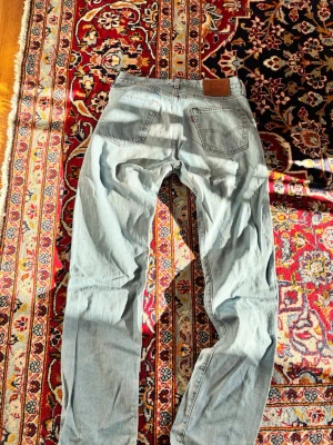 Ljusblåa Levis 501 - Säljer ett par klassiska ljusblå jeans från Levi's med raka ben och femficksmodell. Jeansen har knappgylf och den ikoniska läderpatchen bak i midjan. Perfekta för en avslappnad och tidlös stil.