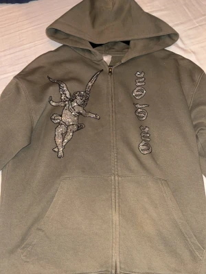 Olivgrön hoodie från one of one - Säljer en olivgrön hoodie med dragkedja och huva. På framsidan finns ett stort motiv av en ängel i strass samt text i gotisk stil. Hoodien har två fickor framtill och är tillverkad i mjukt sweatshirtmaterial. Perfekt för dig som gillar streetwear och unika detaljer. Alla stenar sitter kvar kan lösa fler bilder privat