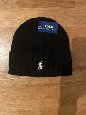 Svart mössa med Polo Ralph Lauren logga - Svart ribbstickad mössa med Polo Ralph Lauren logga broderad framtill. Tjockt uppvik för extra värme och stilren look. Perfekt accessoar för kalla dagar och enkel att matcha med olika outfits.