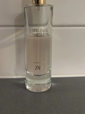 Zara Red Temptation Eau de Parfum - Zara Red Temptation Eau de Parfum i en elegant, genomskinlig glasflaska med gulddetaljer. Doften har en ljusrosa ton och flaskan har en stilren, modern design med raka linjer och tryckt text i svart och guld.