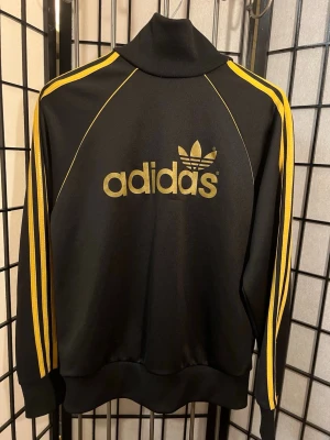 🖤💛 ADIDAS ORIGINALS TRACK JACKET – SVART/GULD – Y2K / AIK VIBE 💛🖤 - Svart & guld = alltid rätt. Riktig adidas Originals med trefoil fram, stor guldlogo bak och diskret adidas-märke i nacken 🔥  Ger sjuk AIK-estetik utan att vara merch. 00s / Y2K / terrace / retro sportswear vibe.  ⚽️🖤💛 Funkar perfekt med: – Svarta jeans – Cargos – TNs / Air Max – Vintage keps  ✨ Detaljer: • Svart bas med gula 3-stripes • Trefoil fram • Stor guld “adidas” på ryggen • Bröst (pit-to-pit): ca 55 cm • Längd: ca 60–61 cm 📦 Skick: bra