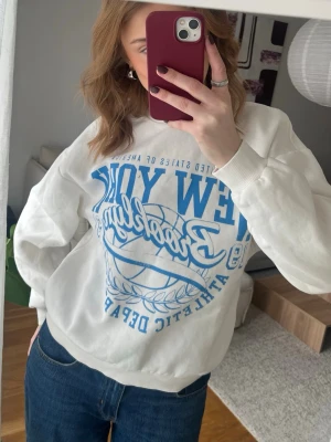 Vit sweatshirt med blått tryck Gina Tricot  - Cool vit sweatshirt med stort blått New York Basketball-tryck på framsidan. Tröjan har rund hals, ribbade muddar och är gjord i mjukt material. Perfekt för en avslappnad och sportig look.