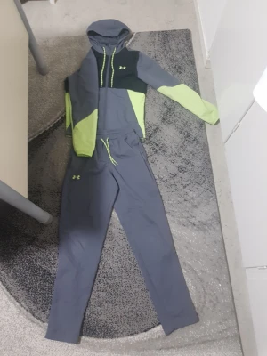 Under Armour tracksuit - Tvådelad tracksuit från Under Armour i grått och limegrönt. Setet består av en hoodie med dragkedja, huva och meshfoder samt matchande byxor med dragsko i midjan. Både jacka och byxor har limegröna detaljer och logga. Perfekt för chill eller träning.