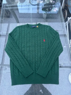 Grön kabelstickad tröja M – Polo Ralph LaurenKlassisk preppy-stil - Tidlös kabelstickad tröja från Polo Ralph Lauren i mörkgrön färg med röd logga på bröstet. Skön, något tjockare kvalitet – perfekt för höst- och vinteroutfits eller lager-på-lager. Passar till jeans, chinos eller skjorta under.  Storlek: M Material: 30% ull, 35% akryl, 35% nylon Tillverkad i: Vietnam Skick: Mycket fint skick, inga fläckar eller hål  ✔ Original ✔ Kabelstickad ✔ Rund hals ✔ Klassisk passform ✔ Trendig grön färg  📦 Skickas snabbt och paketeras noggrant.