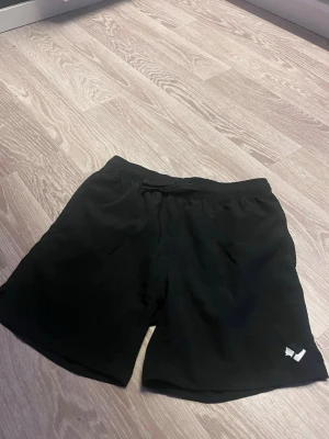 Svarta shorts från 157 Funk - Svarta shorts från 157 Funk med elastisk midja och enkel design. Perfekta för träning eller chill, tillverkade i ett lätt och bekvämt syntetmaterial. Diskret logga nere vid benet ger en clean look.