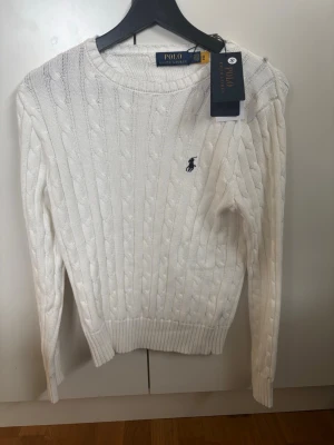 Vit kabelstickad tröja Polo Ralph Lauren - Kabelstickad vit tröja från Polo Ralph Lauren med rund halsringning och broderad marinblå logga på bröstet. Tröjan har ribbade muddar vid ärmslut och nederkant, och är tillverkad i mjukt material som ger en klassisk och clean look. Kan gå ner i pris vid snabbaffär