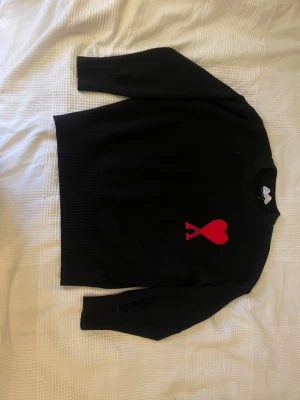 Svart stickad tröja från Ami Paris - Svart stickad tröja från Ami Paris med röd hjärtlogga och bokstaven A på bröstet. Tröjan har rund halsringning, långa ärmar och ribbstickade muddar vid ärmslut och nederkant. Perfekt för dig som gillar stilrena plagg med en twist.