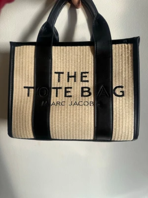 Beige väska från Marc Jacobs - Snygg väska från Marc Jacobs i beige flätad textil med svarta detaljer och handtag. Väskan har texten 'THE TOTE BAG' broderat framtill och är perfekt för dig som vill ha en trendig och rymlig väska till vardagen.