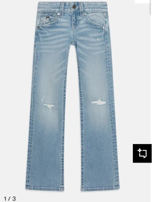 Ljusblå bootcut jeans med slitningar - Säljer ett par ljusblå bootcut jeans med slitna detaljer på låren och klassisk femficksdesign. Jeansen har knapp- och dragkedjegylf samt dekorativa ficklock bak. Perfekta för en avslappnad och trendig look.