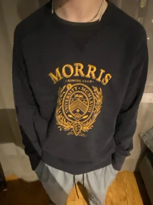 Svart sweatshirt från Morris - Collegeinspirerad svart sweatshirt från Morris med gul tryckt logga och emblem på bröstet. Tröjan har rund halsringning, långa ärmar och ribbade muddar. Perfekt för en avslappnad och stilren look.