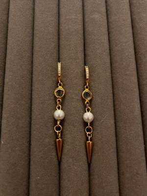 Guldpläterade örhängen med pärlor - Snygga guldpläterade örhängen med vita pärlor och svarta stenar. Hängande design med spetsiga guldfärgade detaljer längst ner. Perfekt accessoar för dig som gillar en mix av klassiskt och edgy stil.