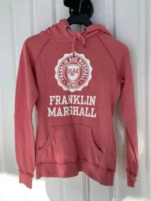 Rosa hoodie från Franklin & Marshall - Snygg rosa hoodie från Franklin & Marshall med vit logga och text framtill. Klassisk känguruficka, dragsko i huvan och ribbade muddar. Har mer att ge, något tunnare i kvaliten och rakare modell, djur och rökfritt hem! Fler bilder vid förfrågan. 