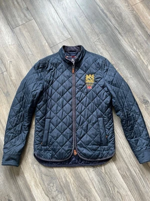 Mörkblå quiltad pufferjacka Morris - Quiltad mörkblå pufferjacka från Morris med guldbroderad logga och brittisk flagga på bröstet. Jackan har ståkrage, dragkedja framtill och två snedställda fickor. Klassisk och stilren design, perfekt för kyliga dagar.