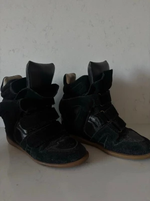Isabel Marant svarta höga sneakers - Säljer mina äkta svarta höga sneakers från Isabel Marant med cool design och kardborreband. Säljer dem då dem inte längre kommer till användning och förtjänar en ny garderob. Skorna har en mix av mocka och läderdetaljer samt gummisula i beige. Perfekta för dig som vill ha en edgy och trendig look. Det finns ett litet slitage på utsidan av skon vid ett spänne. (Skriv för bild)