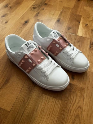 Valentino Rockstud Untitled INSPIRERADE sneakers - Snygga vita Valentino INSPIRERADE skor med rosa band och ikoniska pyramidnitar. Skorna har lågt skaft, klassisk snörning och är tillverkade i slätt läder. Perfekta för dig som vill sticka ut med en lyxig och trendig detalj. Köpta secondhand, endast testade innomhus! Hade behållt om de inte var för små 