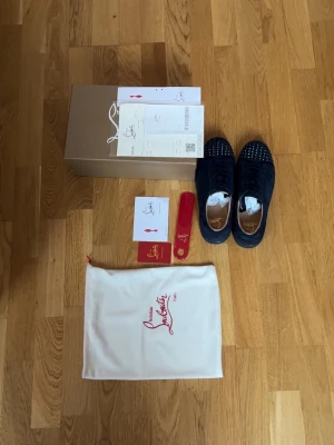 Christian Louboutin sneakers navy blå - Snygga navy blå sneakers från Christian Louboutin med ikoniska röda sulor och svarta nitar på tån. Skorna har lågt skaft och är tillverkade i mocka med läderdetaljer. Perfekta för dig som vill sticka ut med exklusiv design och coola detaljer.