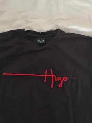 Svart Hugo Boss t-shirt med röd logga - Säljer en svart t-shirt från Hugo Boss med röd broderad Hugo-logga över bröstet. Klassisk rund halsringning och korta ärmar. Tillverkad i mjuk bomull som känns skön mot huden. Perfekt för dig som gillar stilrena plagg med en twist.