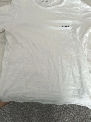 Vit t-shirt från BOSS i bomull - Klassisk vit t-shirt från BOSS med rund halsringning och diskret BOSS-logga på bröstet. Tillverkad i mjuk bomull och har en normal passform. Perfekt basplagg för en clean och stilren look.