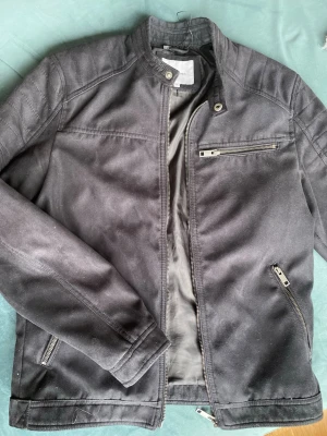 Svart bikerjacka i mocka från Jack & Jones, M - Snygg bikerjacka från Jack & Jones med ståkrage och dragkedja framtill. Jackan har tre fickor med dragkedja och en stilren, minimalistisk design. Den är i storlek M och den är i nyskick.