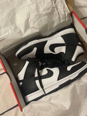 Nike Dunk High Panda sneakers - Svarta och vita Nike Dunk High 'Panda' sneakers med klassisk Swoosh-logga på sidan. Skorna har högt skaft, svart-vit färgkombination och är tillverkade i läder med perforerad tåbox för extra ventilation. Perfekta för dig som gillar streetwear och tidlös stil.