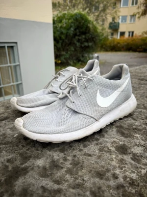 Nike Roshe Run grå sneakers - Säljer ett par grå Nike Roshe Run sneakers med vit sula och klassisk vit swoosh på sidan. Skorna har mesh-överdel för bra ventilation och är riktigt lätta och bekväma. Perfekta för dig som gillar stilrena och sportiga sneakers.