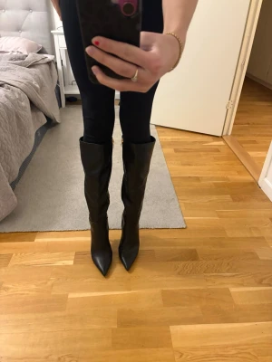 Svarta knähöga boots med spetsig tå - Säljer ett par svarta knähöga stövlar med spetsig tå och slät finish. Stövlarna har en klassisk, stilren design och är tillverkade i ett material som liknar skinn. Perfekta till höstens och vinterns outfits. Klacken är 10cm och det är storlek 39. Aldrig använda då de är lite för små. Pris går att diskutera 