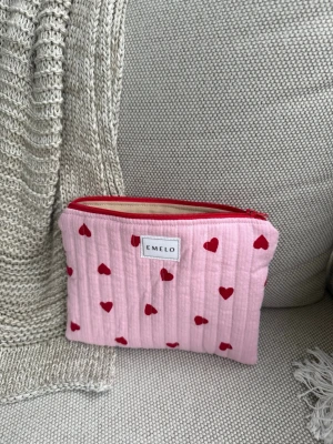 Rosa necessär med röda hjärtan från Emelo - Söt rosa necessär från Emelo med röda hjärtan och röd dragkedja. Tillverkad i mjukt tyg med quiltad känsla och praktisk storlek för småsaker eller smink. Perfekt att ha i väskan för att organisera dina grejer. Mått: 20x15cm