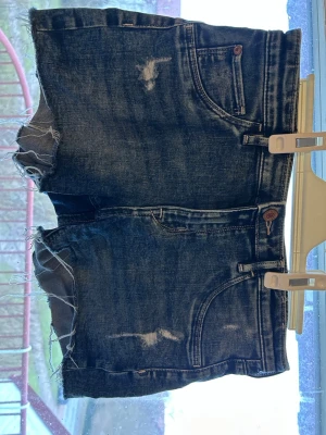 Mörkblå jeansshorts med rå kant - Snygga mörkblå jeansshorts med slitna detaljer och råa kanter. Klassisk femficksmodell med knapp och dragkedja framtill. Perfekta för sommaren och ger en avslappnad vibe.Storlek 170 men skulle säg att de passar som xs/s