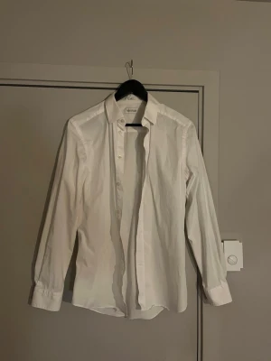 Vit skjorta från These Glory Days - Klassisk vit skjorta från These Glory Days i slim fit-modell. Skjortan har långa ärmar, knappar framtill och är tillverkad i bomull. Perfekt för en stilren och clean look.