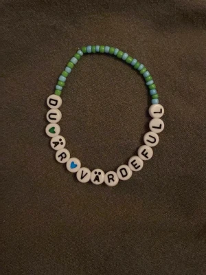 Färgglatt pärlarmband med text - Ett handgjort armband med runda pärlor i grönt, blått och vitt. Armbandet har bokstavspärlor som bildar texten 'DU ÄR VÄRDEFULL' samt små hjärtan och ansikten. Perfekt accessoar för att sprida pepp och glädje.