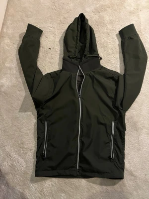 Mörkgrön vindjacka med huva - Snygg mörkgrön vindjacka med dragkedja framtill och två fickor med dragkedjor. Jackan har en justerbar huva och svarta detaljer vid halsen. Perfekt för en vår-sommar dag/kväll och enkel att matcha. Säljer då den är för liten för mig, som är 187