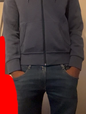 Blå zip hoodie från Polo Ralph Lauren - Mörkblå hoodie med dragkedja från Polo Ralph Lauren. Klassisk design med huva och dragsnören, två stora fickor fram och den ikoniska vita logon broderad på bröstet. Tillverkad i mjukt bomullsmaterial som är skönt att bära.