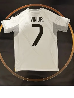 Vit Real Madrid Vini Jr. matchtröja - Säljer en vit Real Madrid matchtröja med VINI JR. och nummer 7 på ryggen. Tröjan har svarta detaljer och klubbens logga under siffran. Perfekt för dig som älskar fotboll och vill stötta ditt favoritlag på planen eller läktaren.