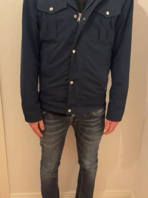 Mörkblå vindjacka med tryckknappar - Snygg mörkblå vindjacka med tryckknappar och dragkedja framtill. Jackan har två bröstfickor med lock och lång ärm. Perfekt för dig som vill ha en stilren och enkel jacka till vardagslooken.skriv om undringar 