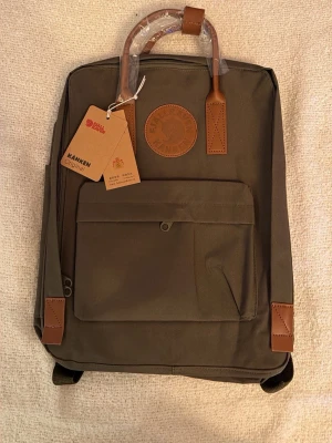 Olivgrön Kånken ryggsäck Fjällräven - Snygg olivgrön Kånken Original ryggsäck från Fjällräven med detaljer i brunt läder. Klassisk fyrkantig form, stort huvudfack, framficka med dragkedja och bärhandtag i läder. Perfekt för skolan eller utflykter.