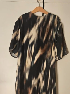 Mönstrad kortklänning i svart och beige - Cool kortklänning med abstrakt mönster i svart, beige och bruna toner. Klänningen har korta ärmar och rak passform, perfekt för dig som gillar en avslappnad men ändå trendig stil. Materialet känns lätt och smidigt mot huden.