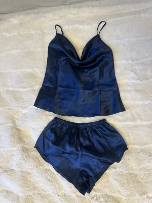 Mörkblått satinsovset med linne och shorts - Snyggt tvådelat sovset i mörkblå satin med ett linne med tunna axelband och draperad urringning samt matchande shorts med resår i midjan. Perfekt för en lyxig känsla hemma. Materialet är glansigt och mjukt mot huden.