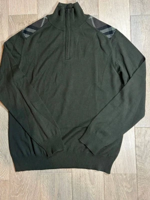 Mörkgrön Burberry tröja - Mörkgrön stickad tröja med halv dragkedja och hög krage. Tröjan har långa ärmar, ribbade muddar och axelpartier med rutigt mönster i svart, vitt och grått. Perfekt för dig som gillar stilrena detaljer.